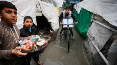 طفل غزّي يقود دراجة هوائية حاملا بين ذراعيه صندوق مساعدات عليه شعار وكالة الأونروا وسط الخيم الغارقة في المطر.