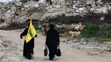 تسير النساء بالقرب من المباني المدمرة، إحداها تحمل علم حزب الله في قرية كفركلا بجنوب لبنان
